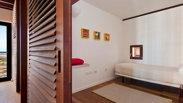 A bedroom with a bed, a red pillow, a wall-mounted shelf, and three framed pictures. - Casas Bioclimáticas Iter El Cubo (Granadilla de Abona)