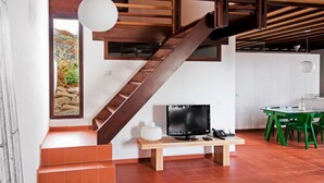 A modern interior with a wooden staircase, a flat-screen TV on a wooden stand, a dining area with green chairs, and a view of a rocky landscape through a window. - Casas Bioclimáticas Iter El Cubo (Granadilla de Abona)