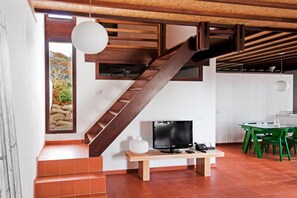 Een modern interieur met een houten trap, een flatscreen televisie op een houten standaard, een eethoek met groene stoelen en uitzicht op een rotsachtig landschap door een raam.