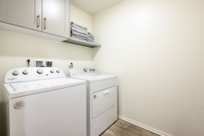 A laundry room with two Whirlpool washers and dryers, a shelf above with folded towels, and a white wall.