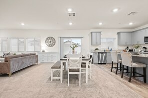 Une cuisine moderne avec des meubles blancs, un coin repas doté d’une table et de chaises blanches, et un salon avec un canapé gris.