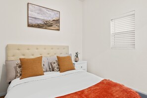 A bedroom with a bed, pillows, a nightstand, a vase, and a painting on the wall.