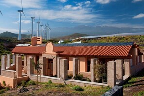 Ein Gebäude mit rotem Ziegeldach, umgeben von Windkraftanlagen und Solaranlagen, vor einer Bergkulisse.