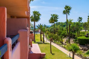 A balcony with a view of a landscaped area with palm trees and a pool. - Spacious Apartment in Casares del Sol (Near the Beach and Finca Cortesin) (Casares)