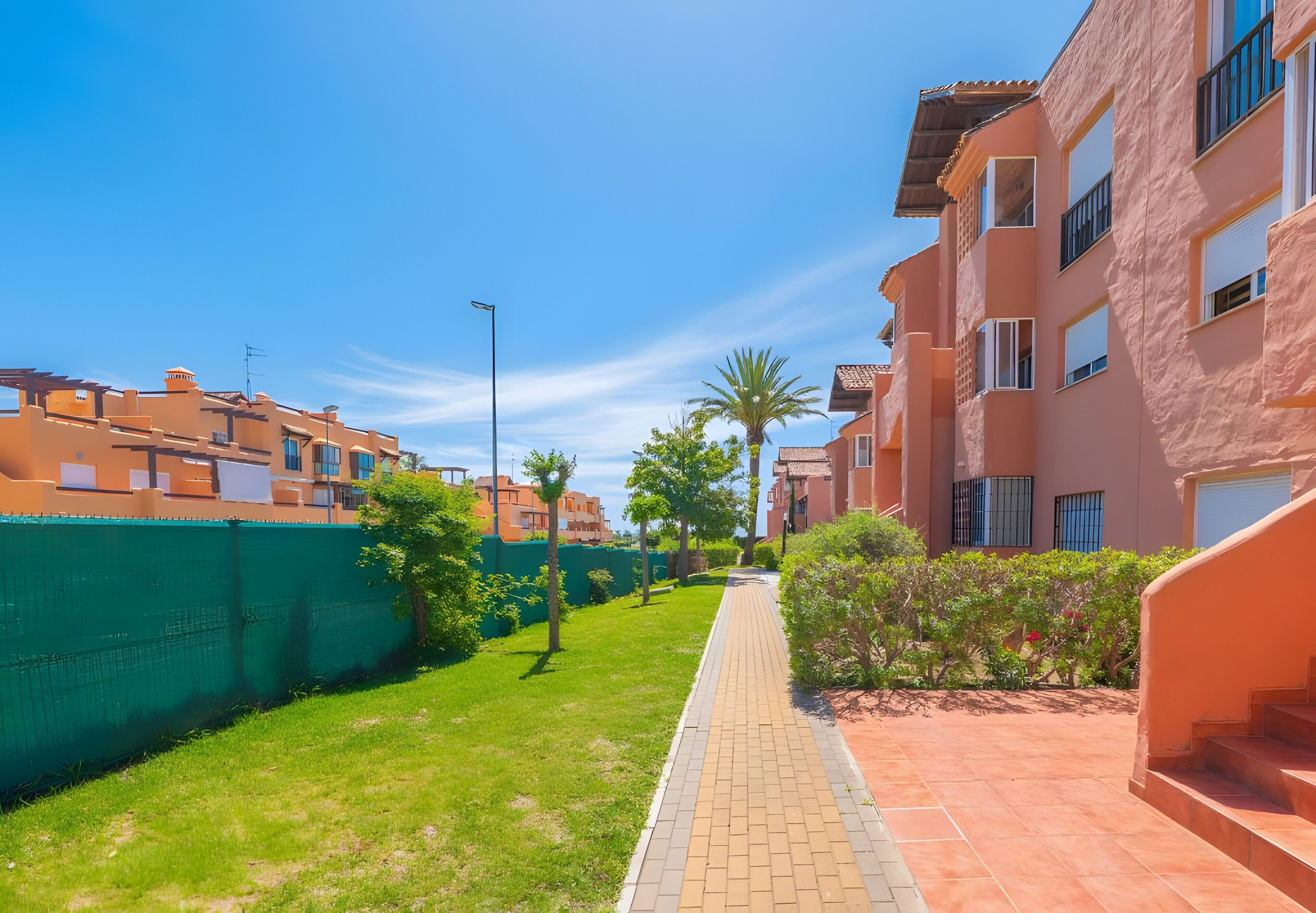 A residential area with a paved walkway, green fencing, and buildings with balconies.