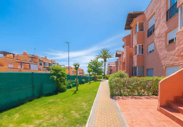 A residential area with a paved walkway, green fencing, and buildings with balconies.