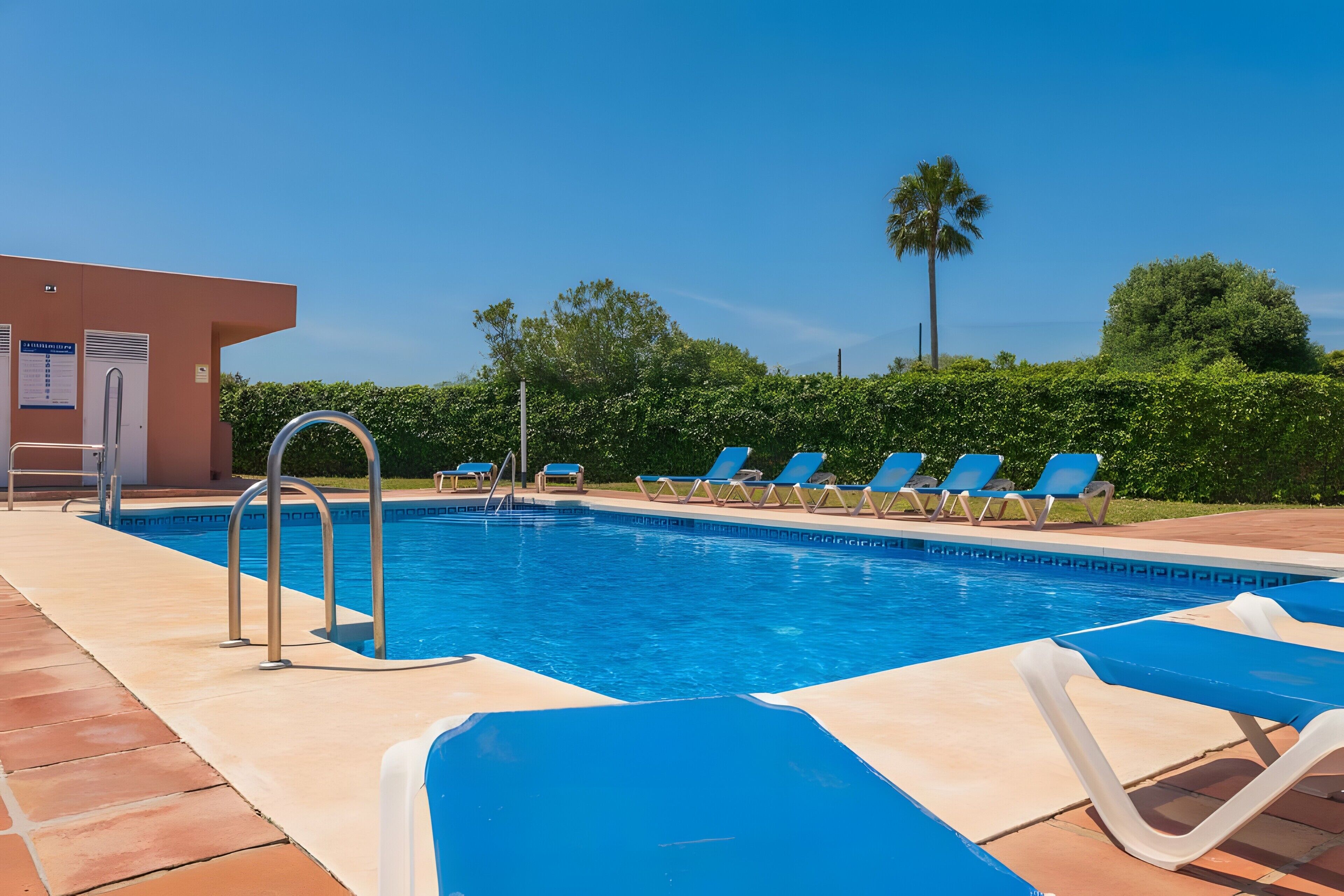 A swimming pool with blue water, surrounded by lounge chairs and a building with a door and windows.