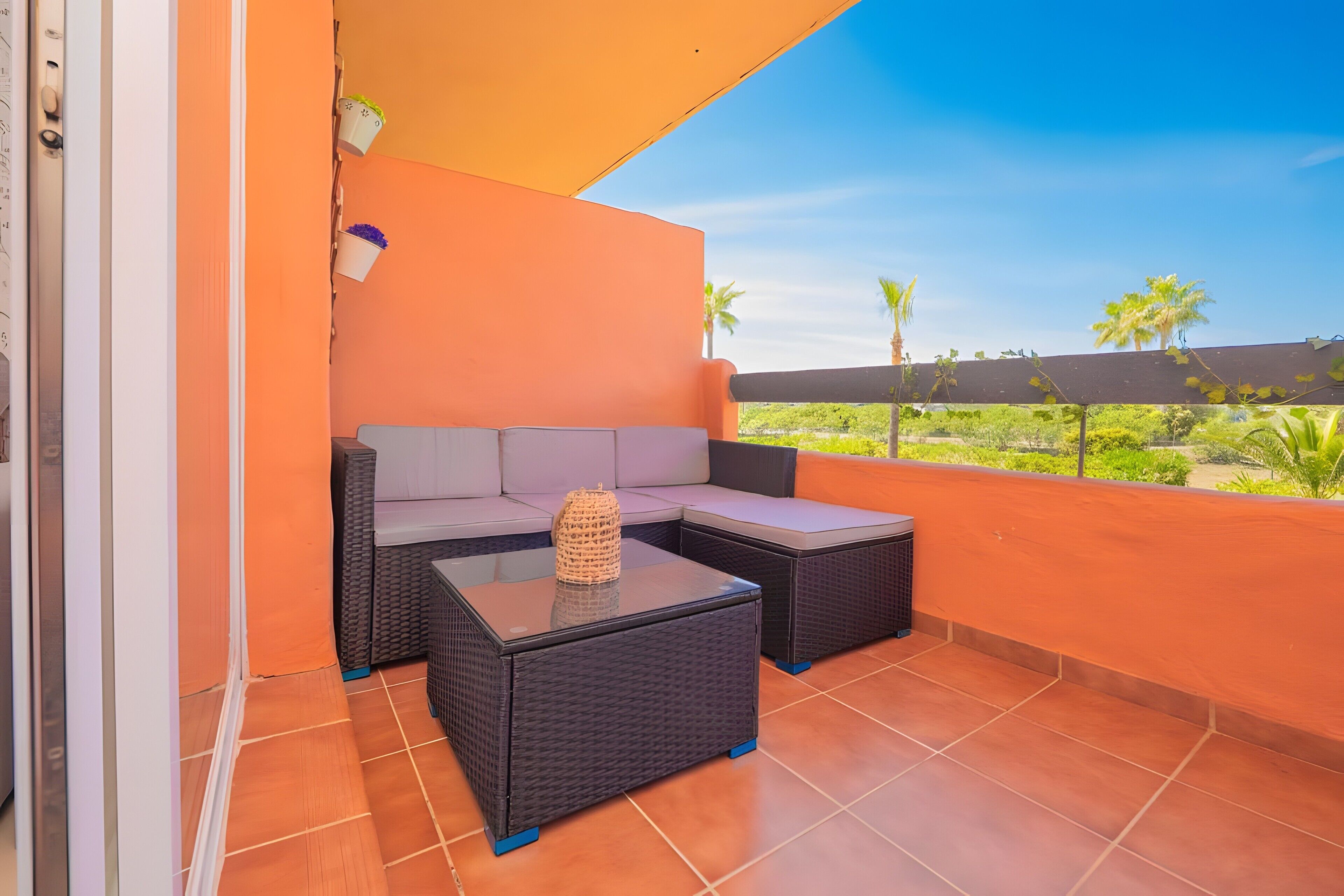 Ein Balkon mit einer Couch und zwei Tischen, Fliesenboden und Blick auf Grünpflanzen und Palmen.