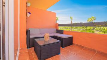 Ein Balkon mit einer Couch und zwei Tischen, Fliesenboden und Blick auf Grünpflanzen und Palmen.