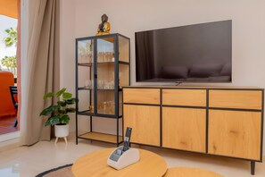 A modern living room with a flat-screen TV, a glass-fronted cabinet with wine glasses, a potted plant, and a round coffee table.