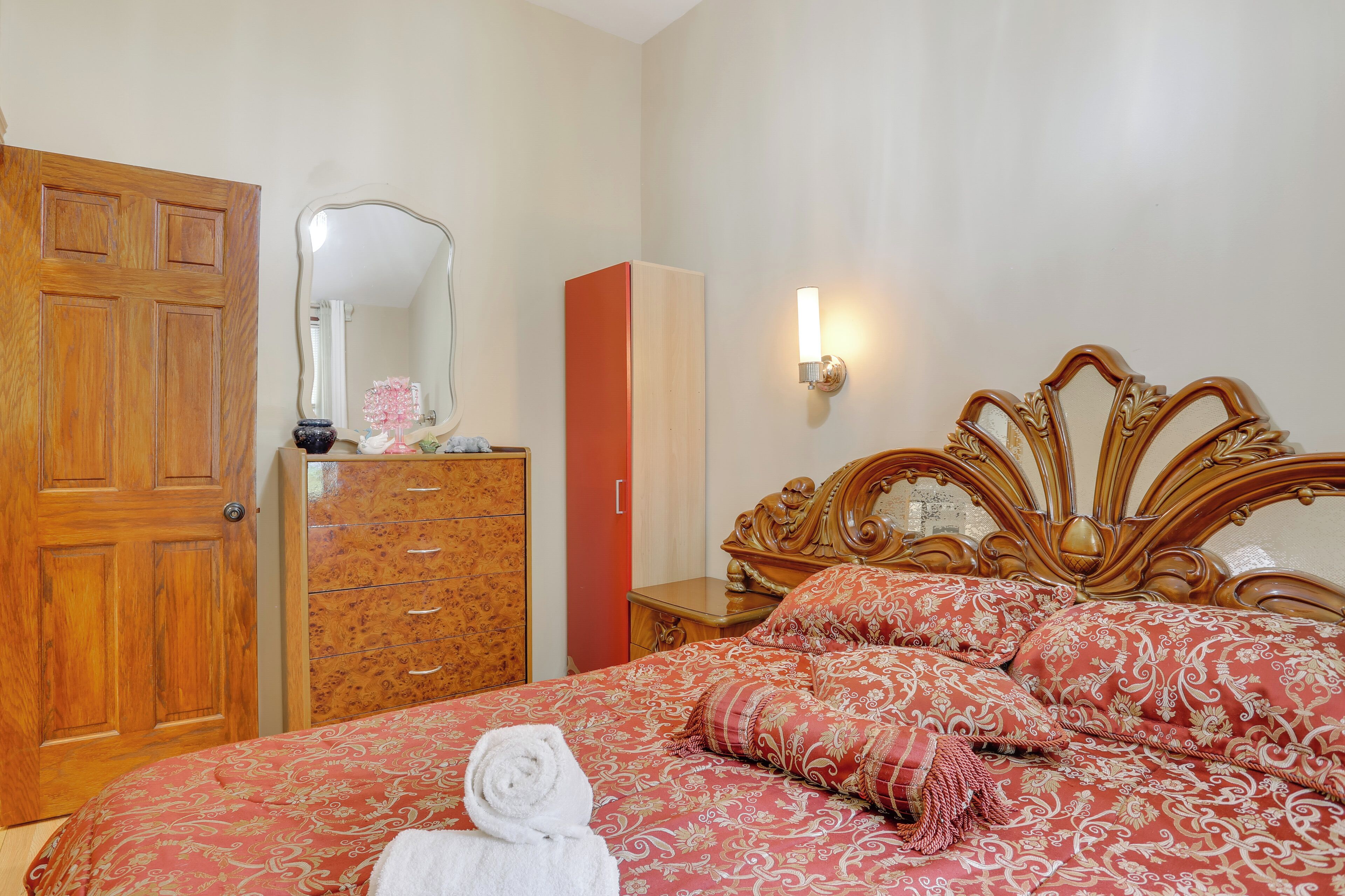 A bedroom with a wooden door, a dresser with drawers, a bed with a floral bedspread and pillows, a nightstand with a lamp, and a mirror on the wall.