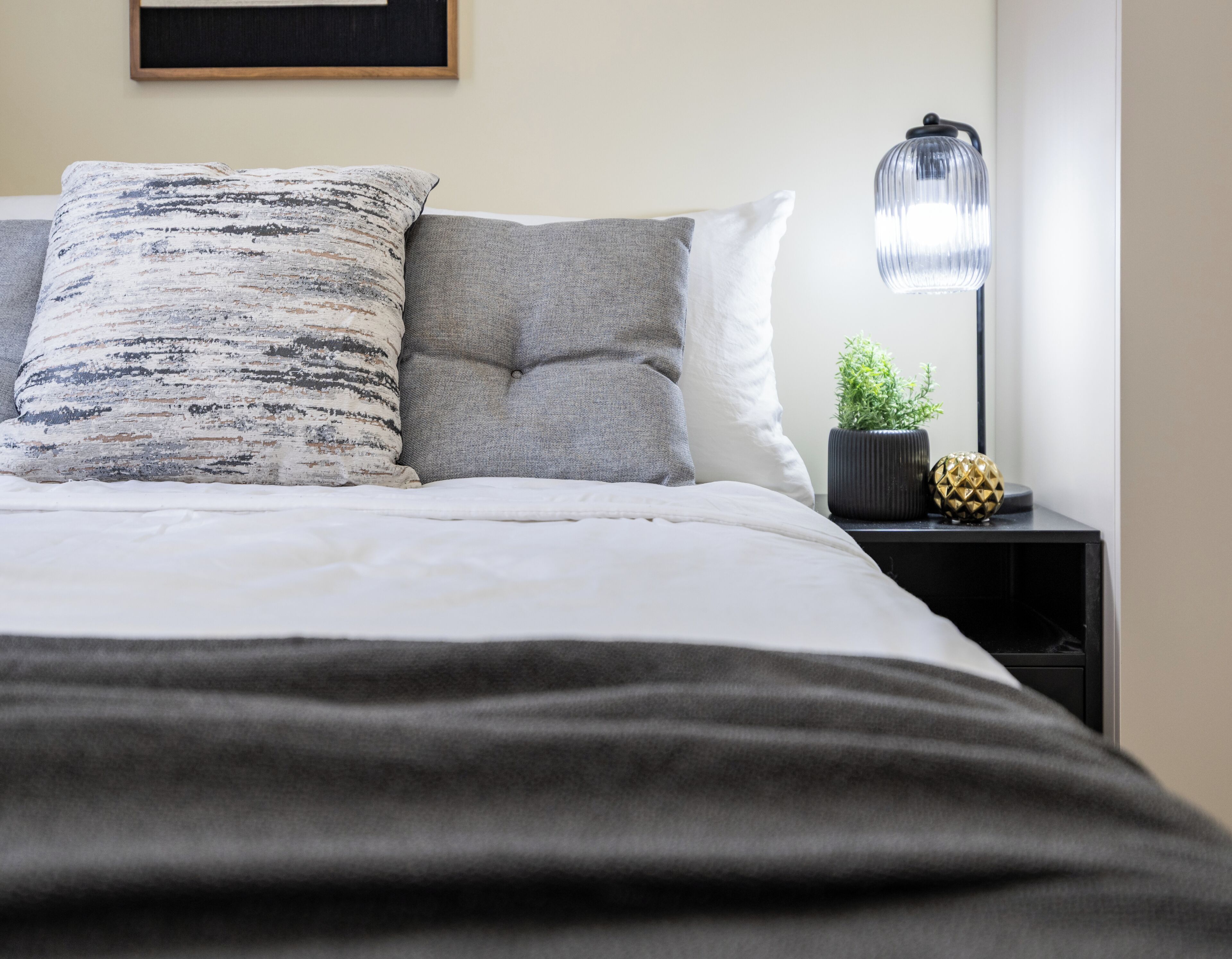 A neatly made bed with a grey blanket, two pillows, a small potted plant, and a lamp on a bedside table.