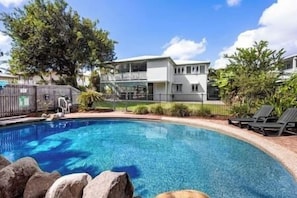 Ein Schwimmbecken mit klarem, blauem Wasser, umgeben von einem Zaun, und ein Gebäude mit einem Balkon im Hintergrund.