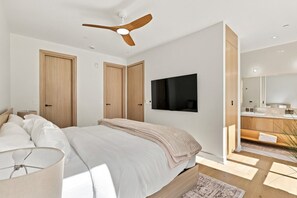 A modern bedroom with a bed, a ceiling fan, a TV, and a bathroom with a mirror and sink. - Modern Mont-Tremblant Getaway -Ostrya (Mont-Tremblant)
