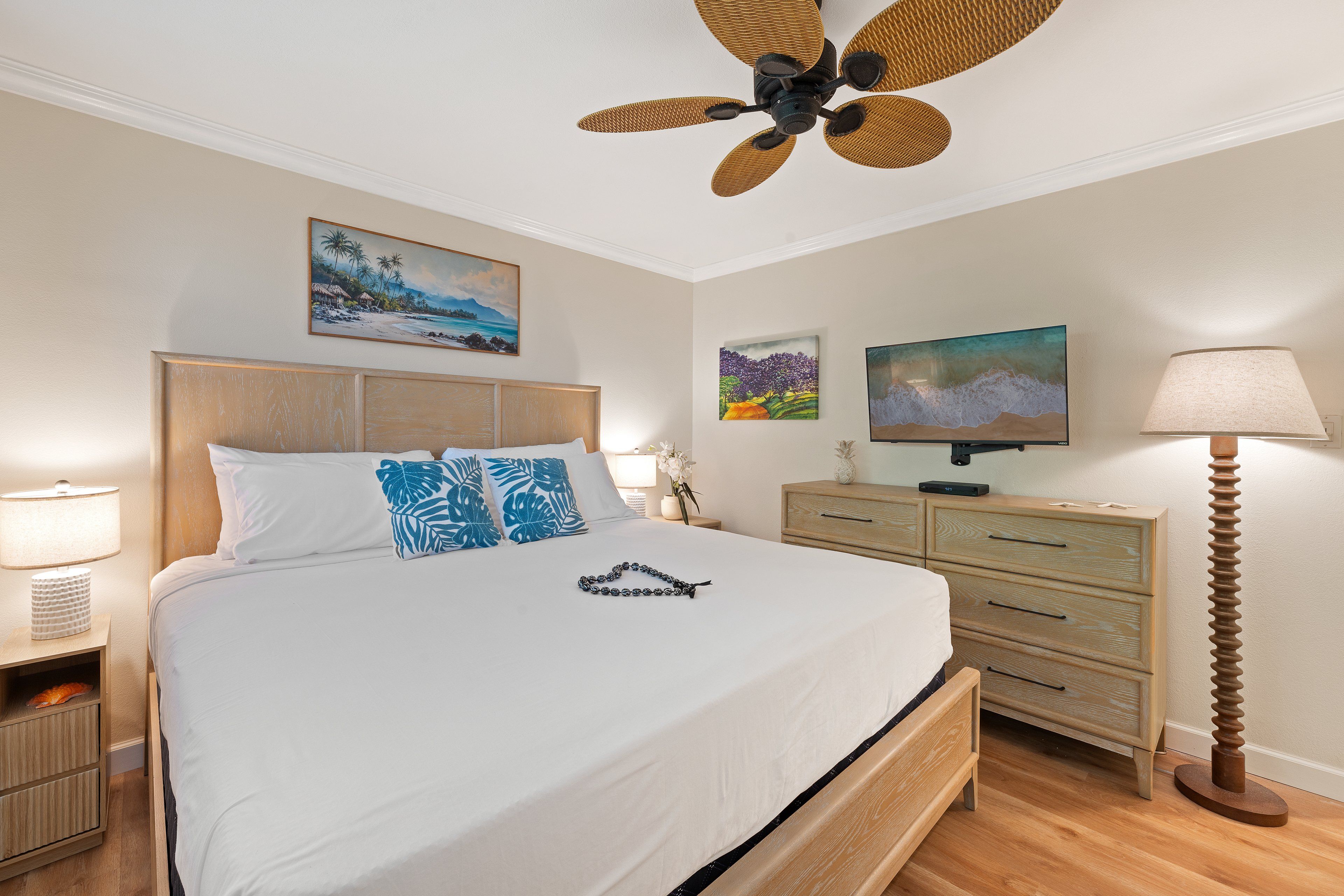 Una camera d'albergo con un letto, una testiera in legno, un comodino con una lampada, una cassettiera, una televisione e una ventola a soffitto.