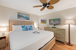 A hotel room with a bed, a wooden headboard, a nightstand with a lamp, a dresser, a television, and a ceiling fan. - Garden View Condo Just Steps from the Ocean -Maui Sands Seaside 717 (Lahaina)
