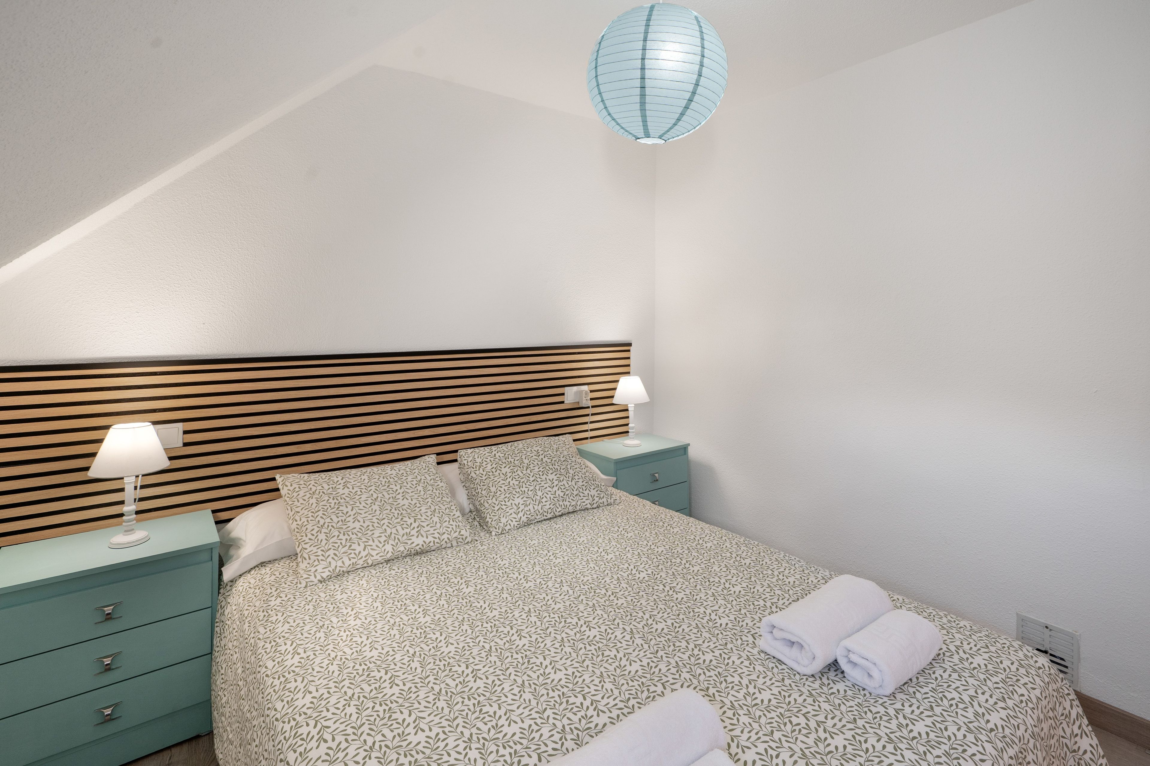 Una cama bien tendida con colcha estampada, dos mesitas de noche con lámparas, cabecera de madera con listones y una lámpara colgante.
