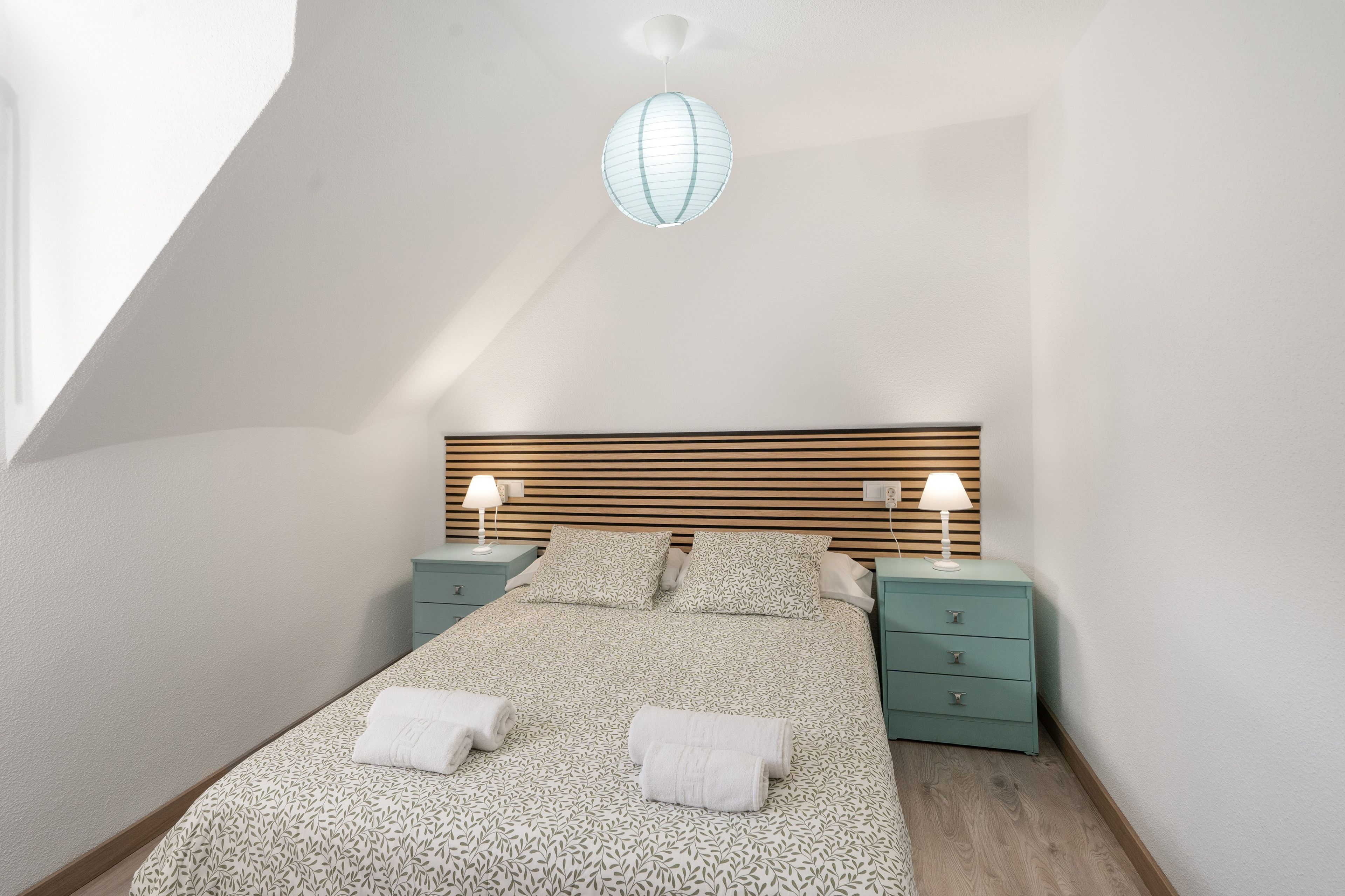 Ein Schlafzimmer mit geneigter Decke, einem Bett mit blattgemustertem Überwurf, zwei Nachttischen mit Lampen und einer Hängeleuchte.