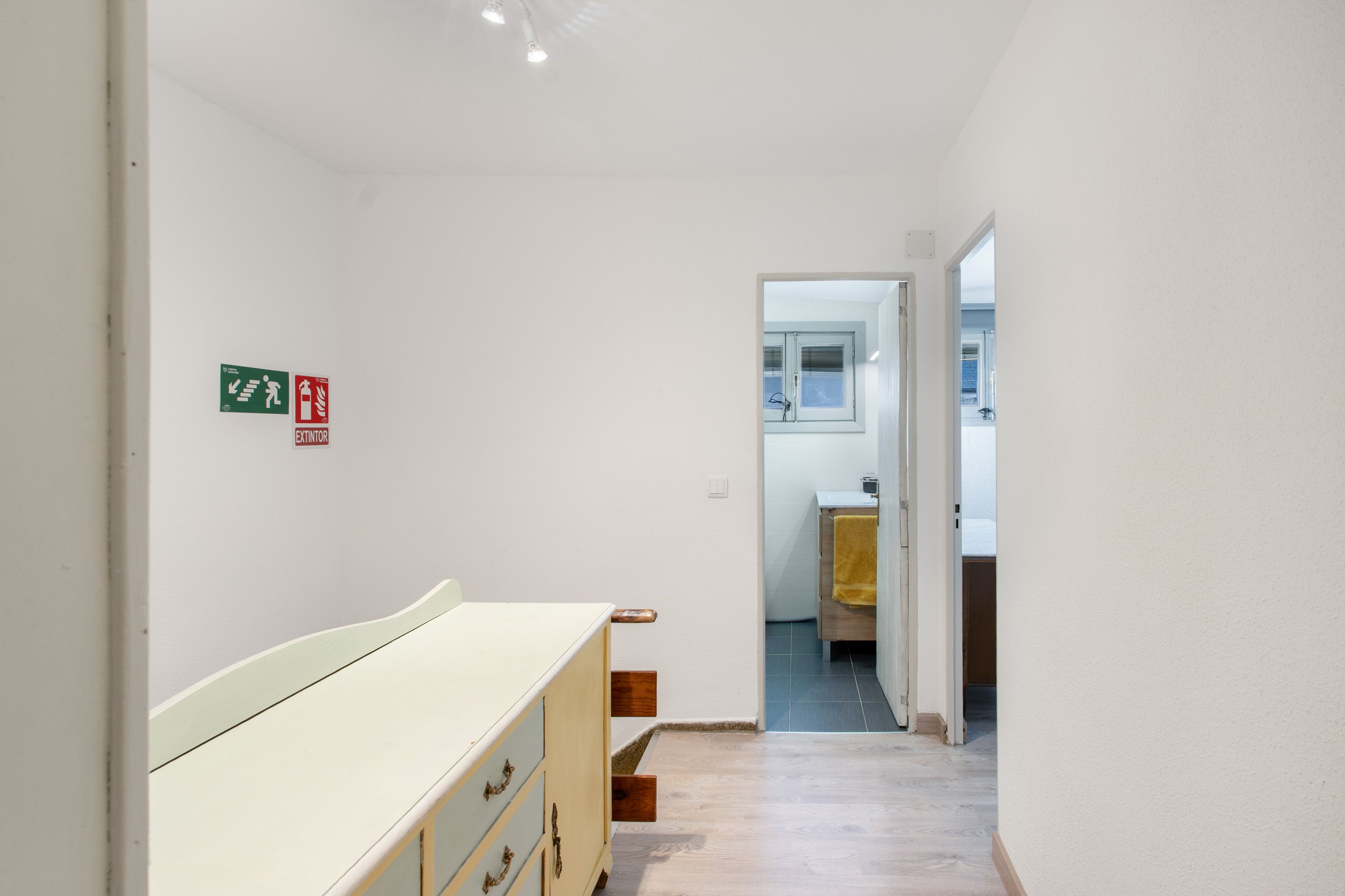 Un pasillo con un aparador blanco, un letrero de salida de incendios y un baño visible a través de una puerta abierta.
