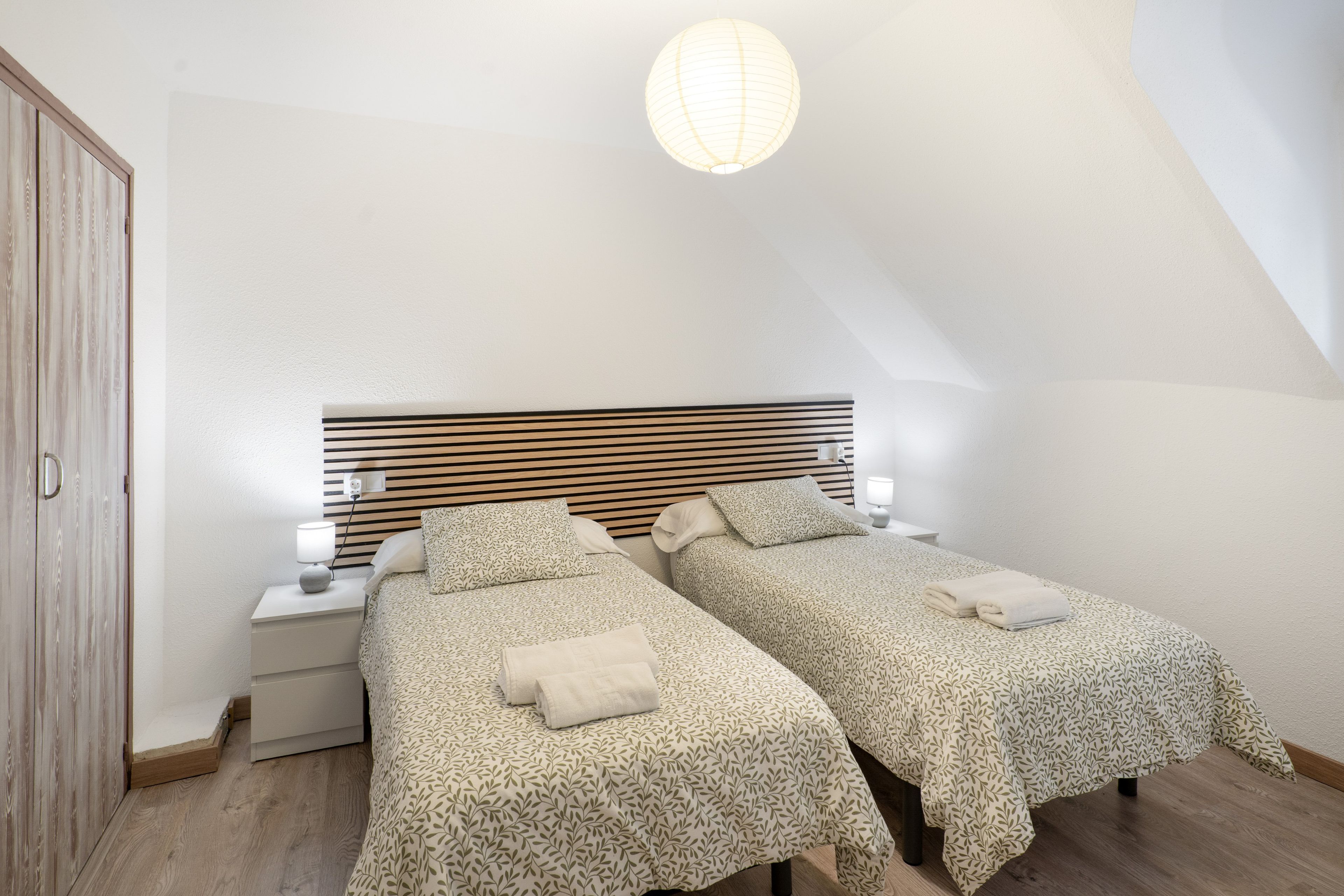 A bedroom with two beds, a wooden headboard, bedside lamps, and a hanging light fixture.