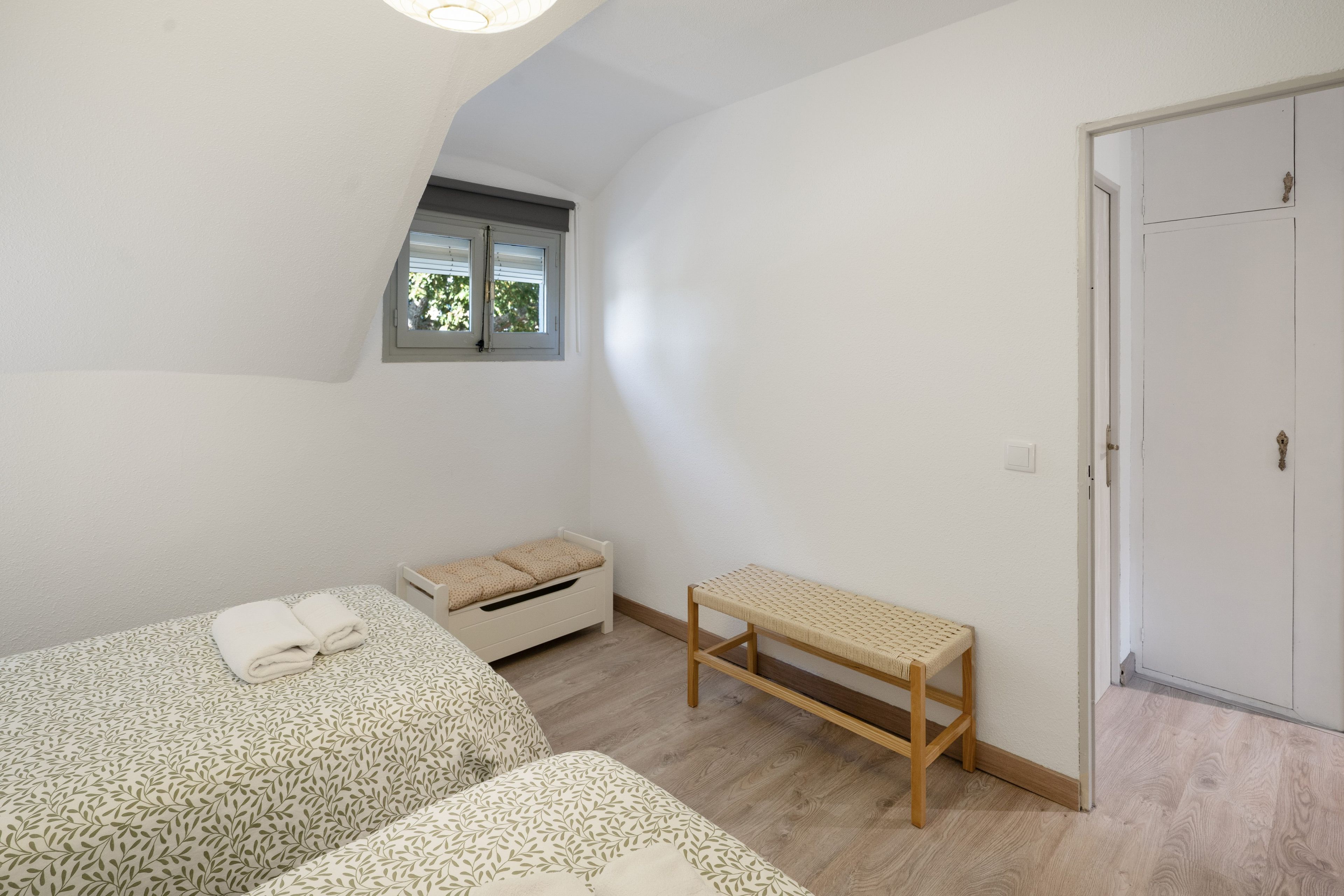 Una habitación pequeña y luminosa con dos camas, un banco, un ventanal y una puerta.