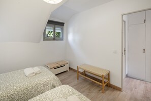 A small, well-lit room with two beds, a bench, a window, and a door.