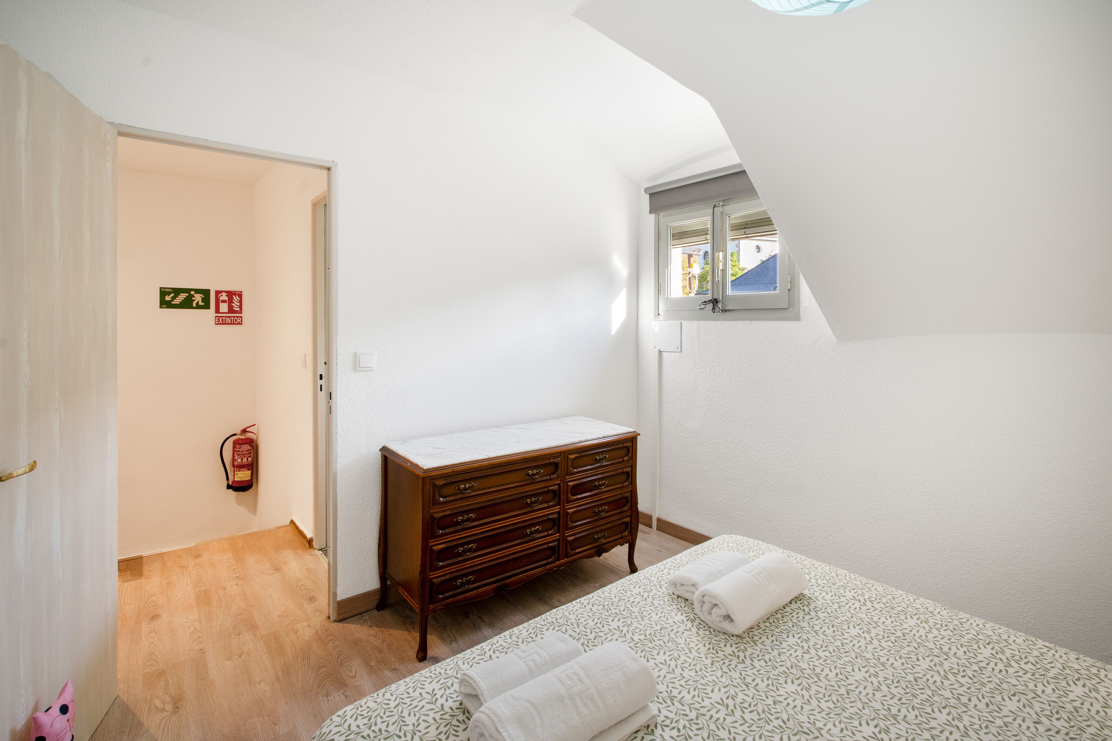 Ein Schlafzimmer mit einem Holzkasten, einem Bett mit gemustertem Überwurf, einem Fenster mit Ausblick und einem Notausgangsschild.