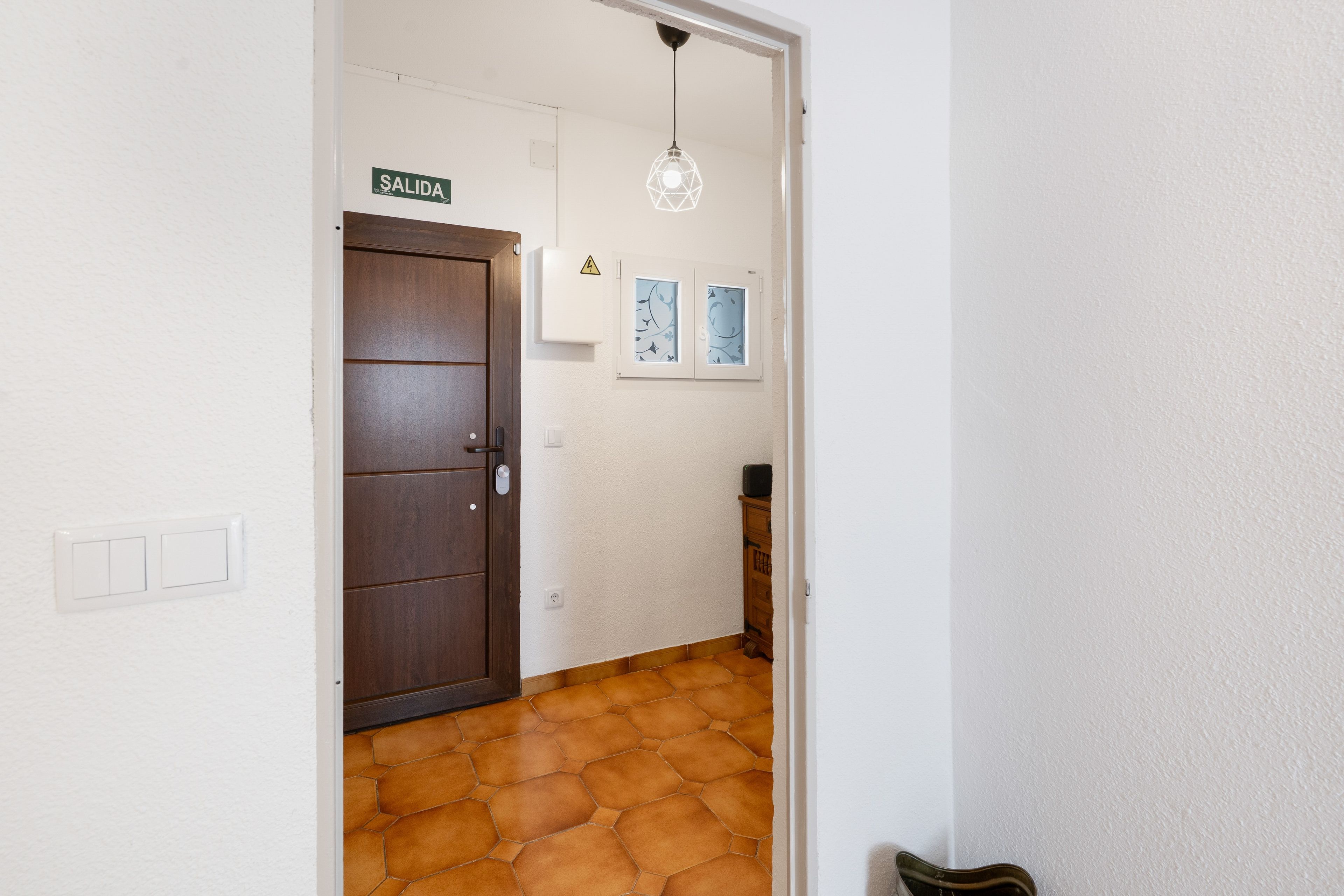 Un pasillo con una puerta marrón, una pared blanca, piso de baldosas, una lámpara en el techo y un pequeño armario.