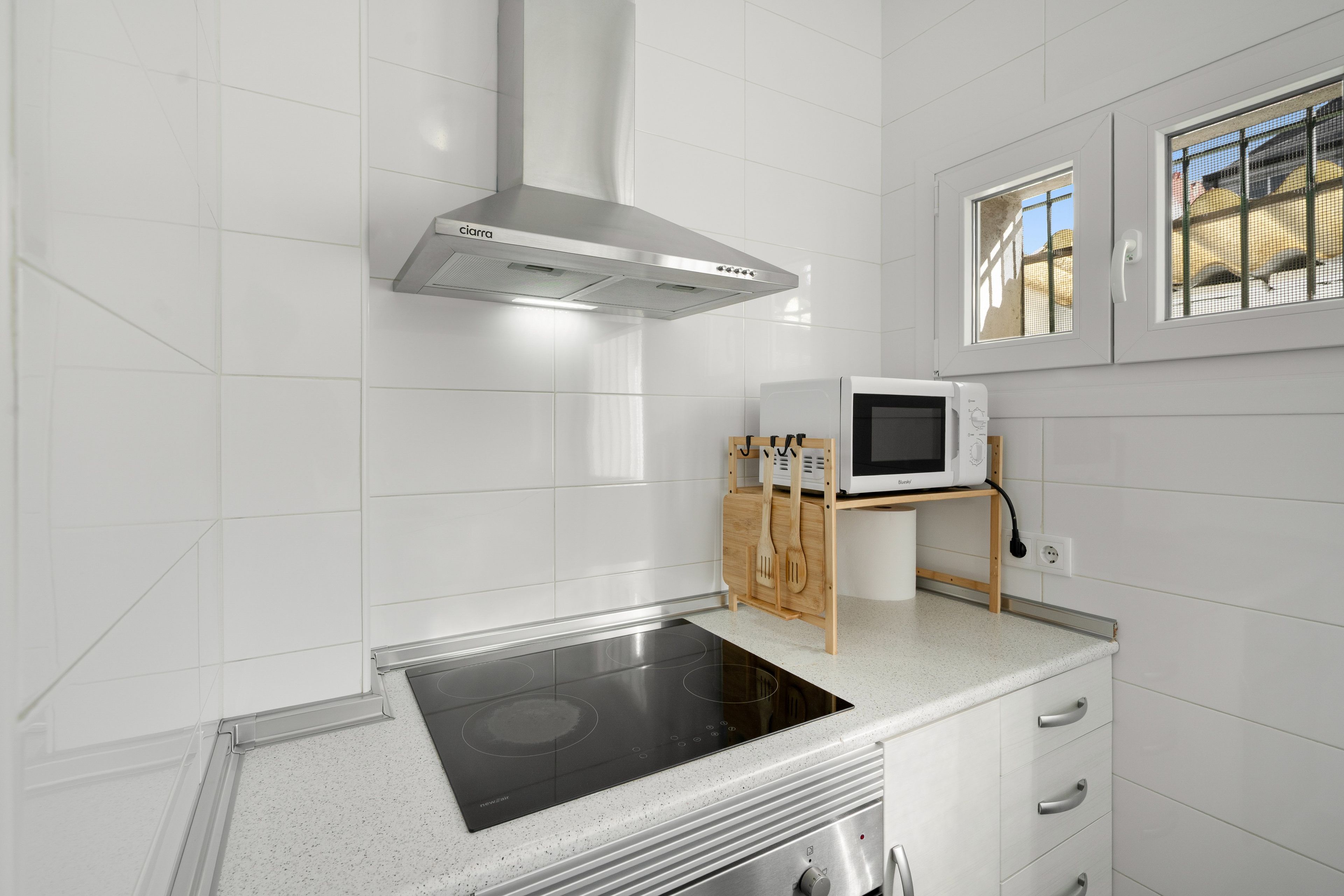 Una cocina moderna con azulejos blancos, una campana extractora de acero inoxidable, un microondas sobre un soporte de madera y una placa de inducción integrada.