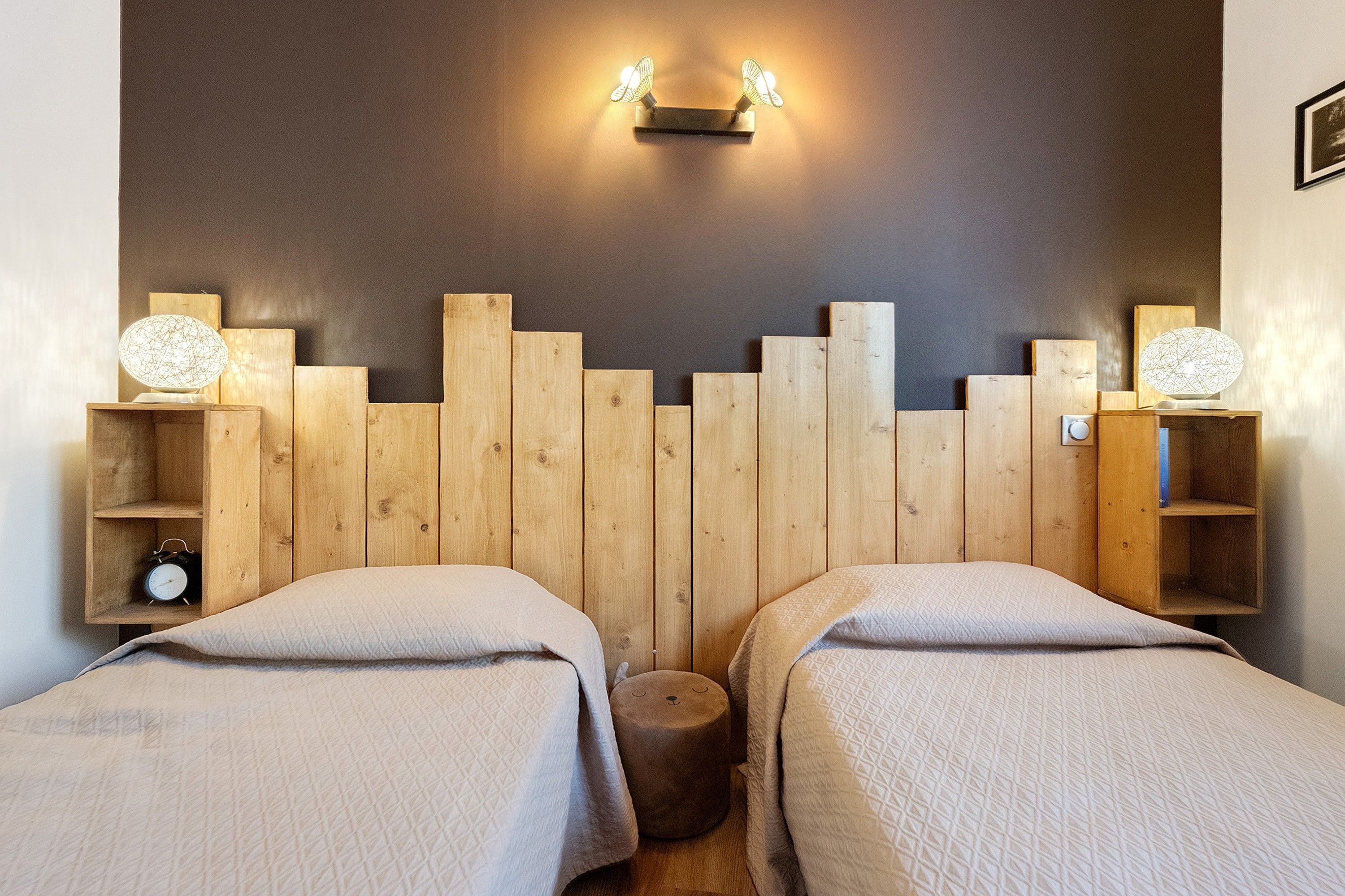A room with two beds, a wooden headboard, bedside tables with lamps, and a wall-mounted light fixture.