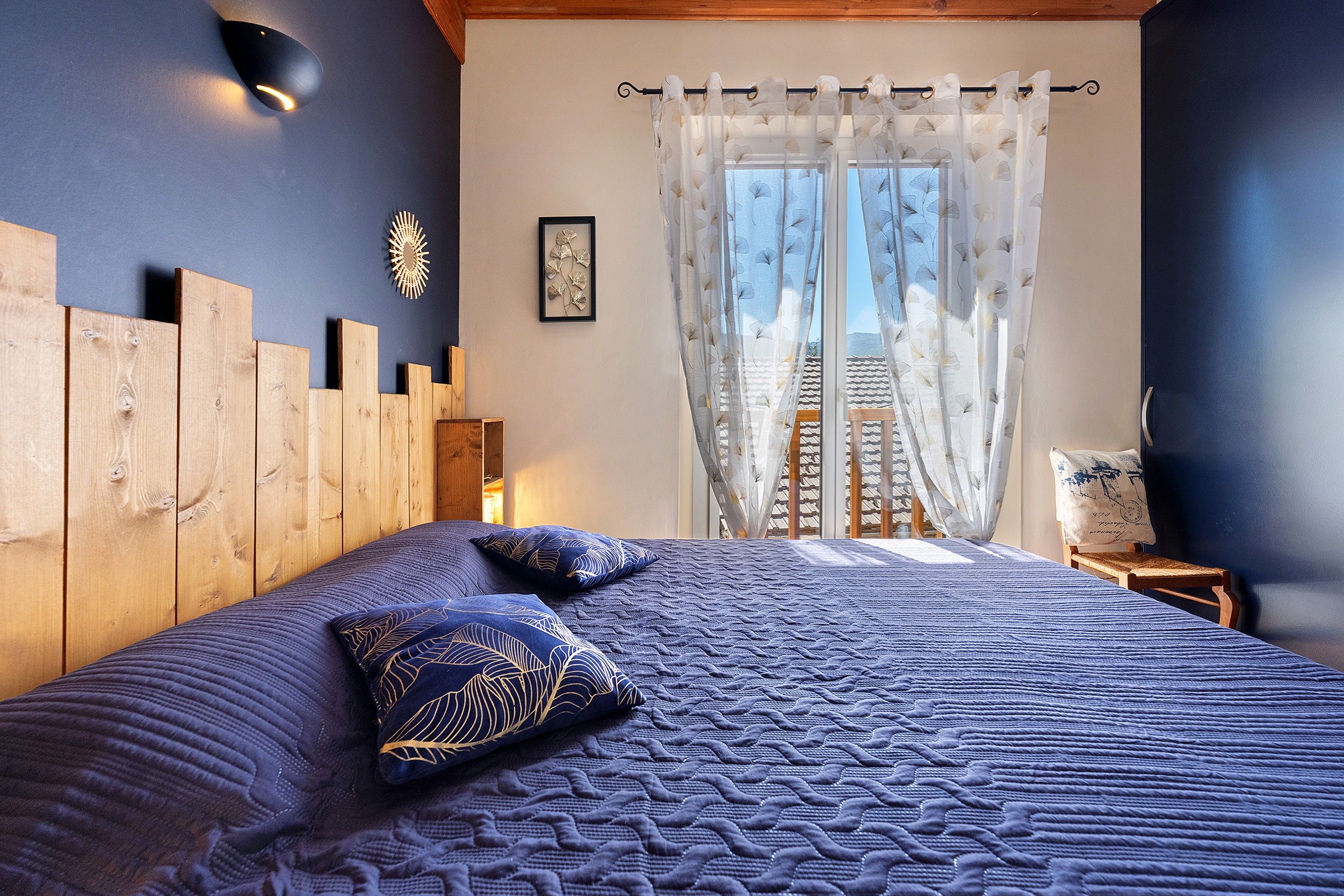 Ein Schlafzimmer mit einem großen Bett, einem hölzernen Kopfteil und Blick auf einen Balkon durch leichte Vorhänge.