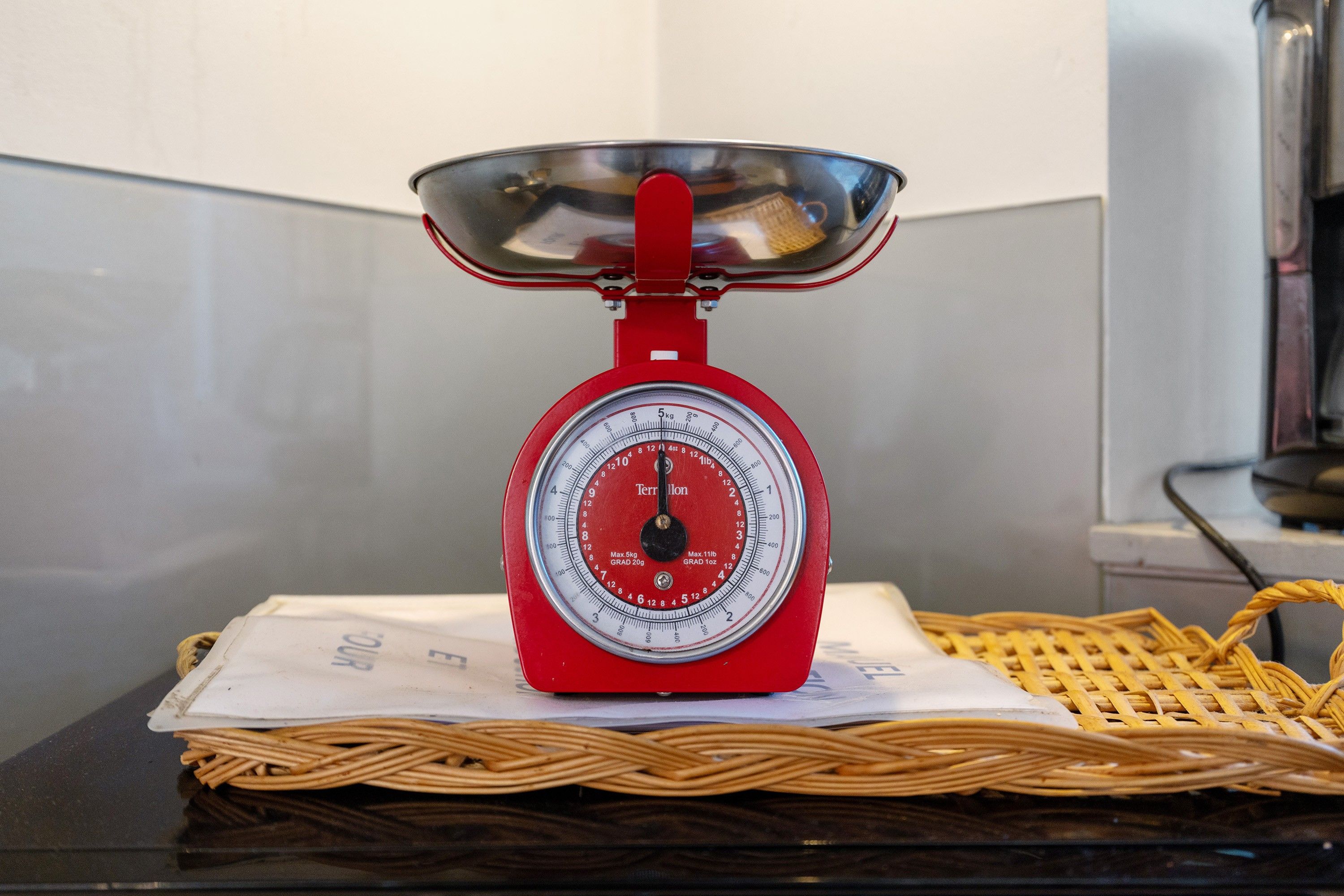 A red kitchen scale with a stainless steel bowl on top, placed on a woven mat over a dark surface.