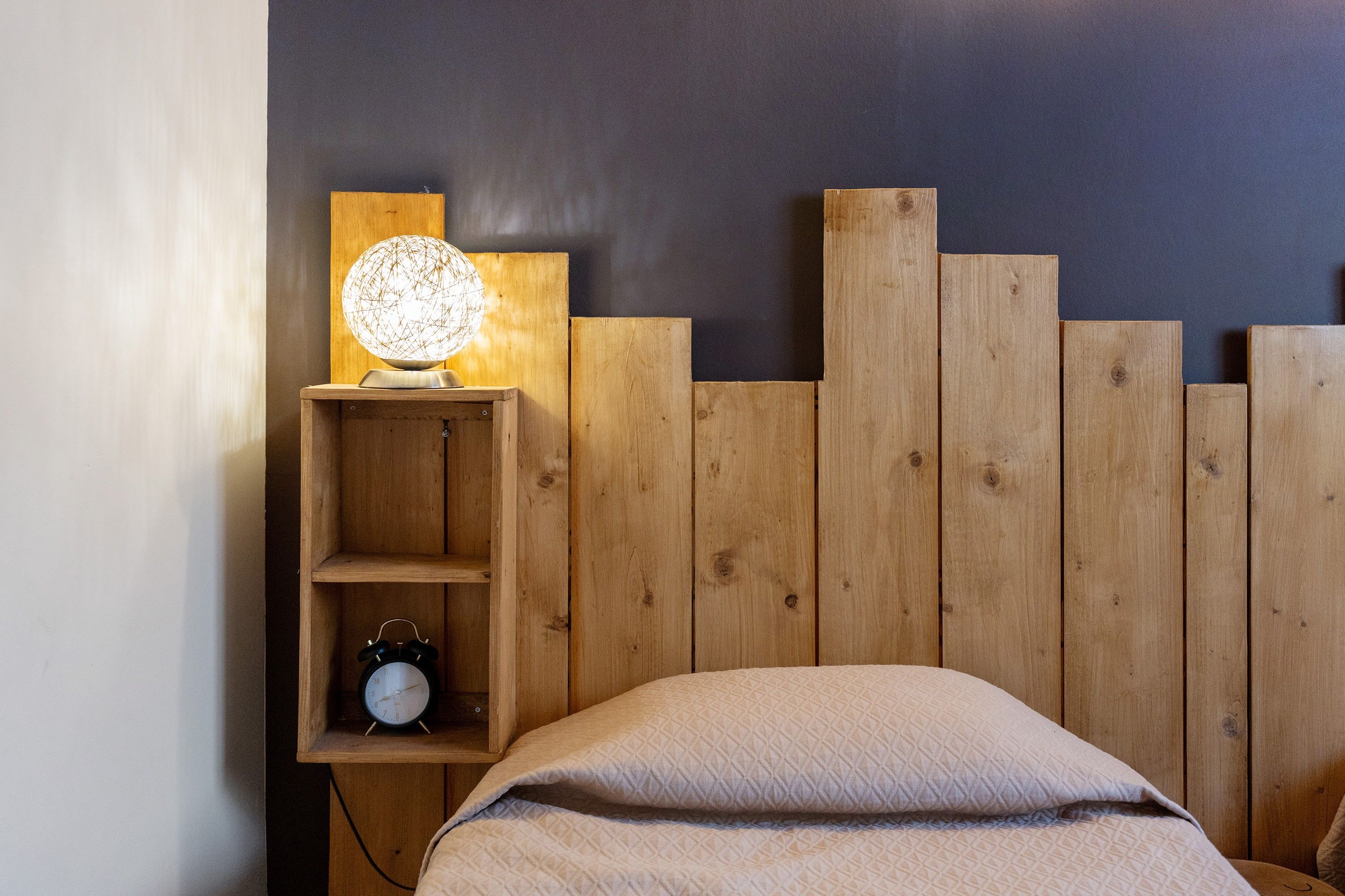 Une chambre à coucher avec une tête de lit en bois, une table de chevet avec une lampe et un réveil, et un lit recouvert d’une couverture matelassée.