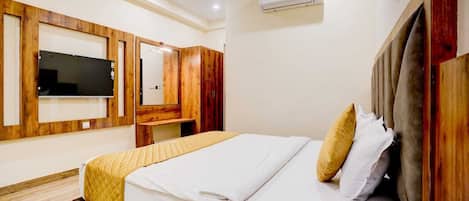 A hotel room with a bed, a TV mounted on the wall, a wooden wardrobe, and an air conditioning unit.
