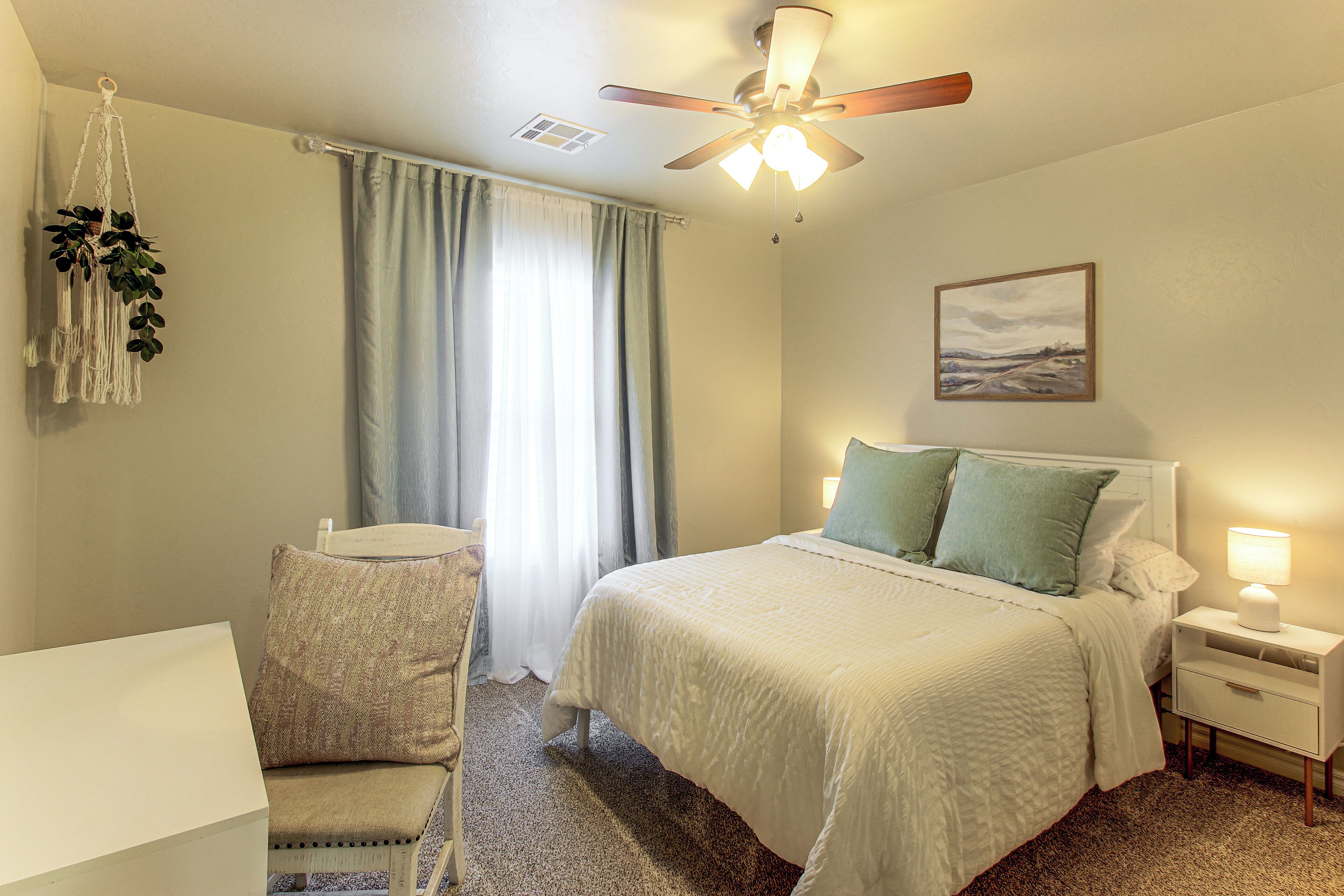 A hotel room with a bed, a desk, a chair, a nightstand, a lamp, a ceiling fan, and a painting on the wall.