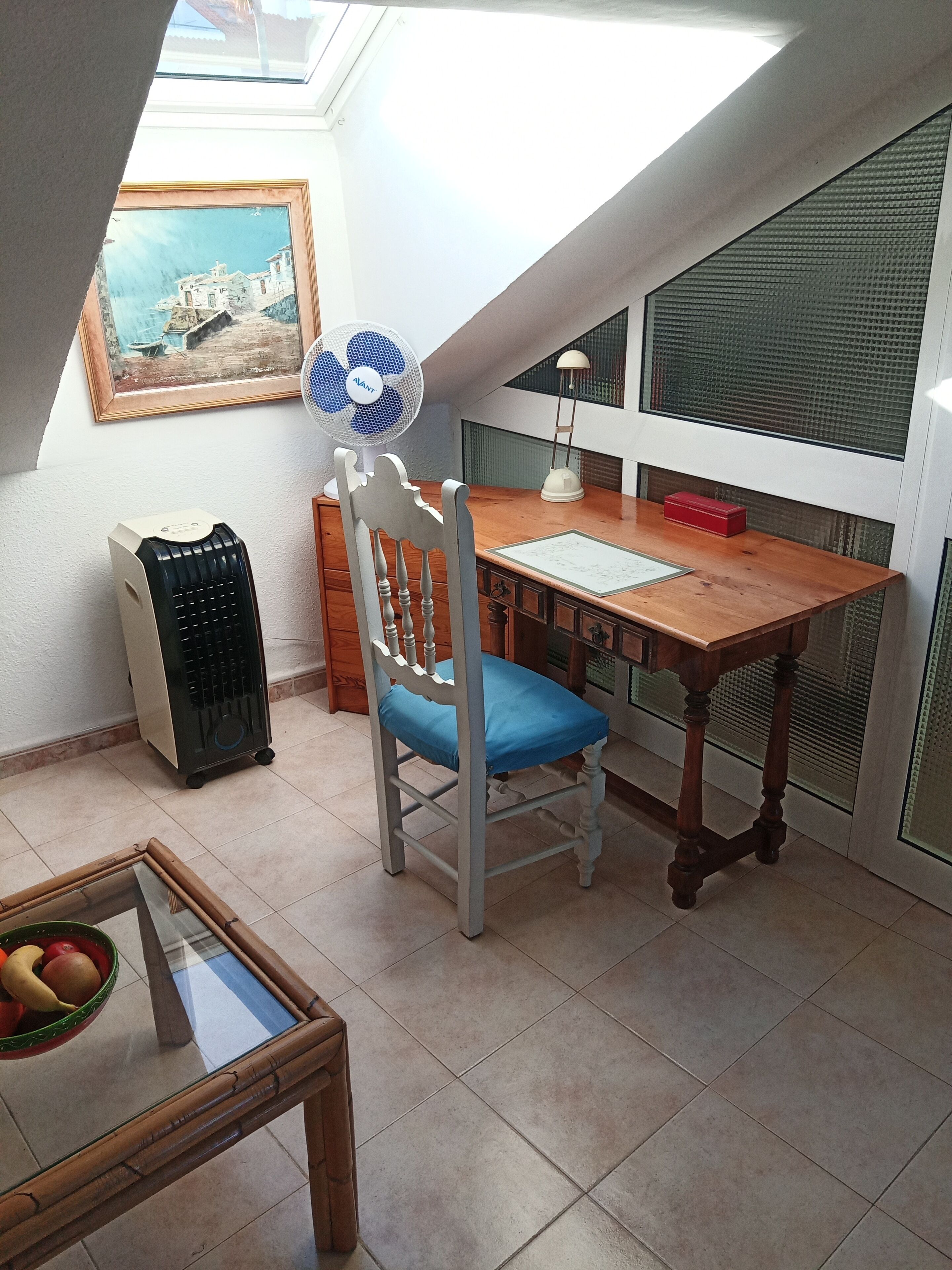 Una habitación con techo inclinado, una mesa de madera, una silla, un ventilador, una lámpara, un radiador y un cuadro en la pared.