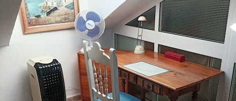 Una habitación con techo inclinado, una mesa de madera, una silla, un ventilador, una lámpara, un radiador y un cuadro en la pared.