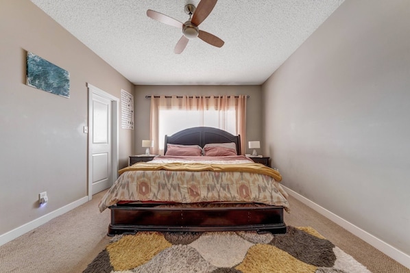 A bedroom with a bed, a ceiling fan, a window with curtains, a door, and a wall-mounted artwork. - Shalom Casablanca Home (Edmonton)