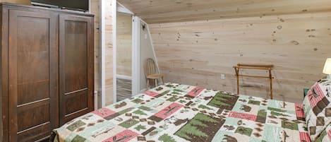 A bedroom with a wooden ceiling, a bed with a patterned quilt, a wooden wardrobe, and a chair.