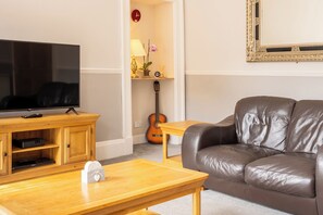 A living room with a television on a wooden stand, a leather sofa, a wooden coffee table, a guitar, and a wall-mounted clock. - Incredibly Located 3BDR Flat in Edinburgh (Edinburgh)