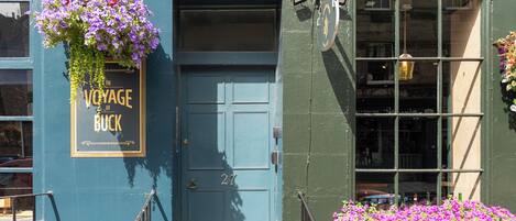 A building with a blue door marked with the number 27, a hanging flower basket, and a window with a view of the street.