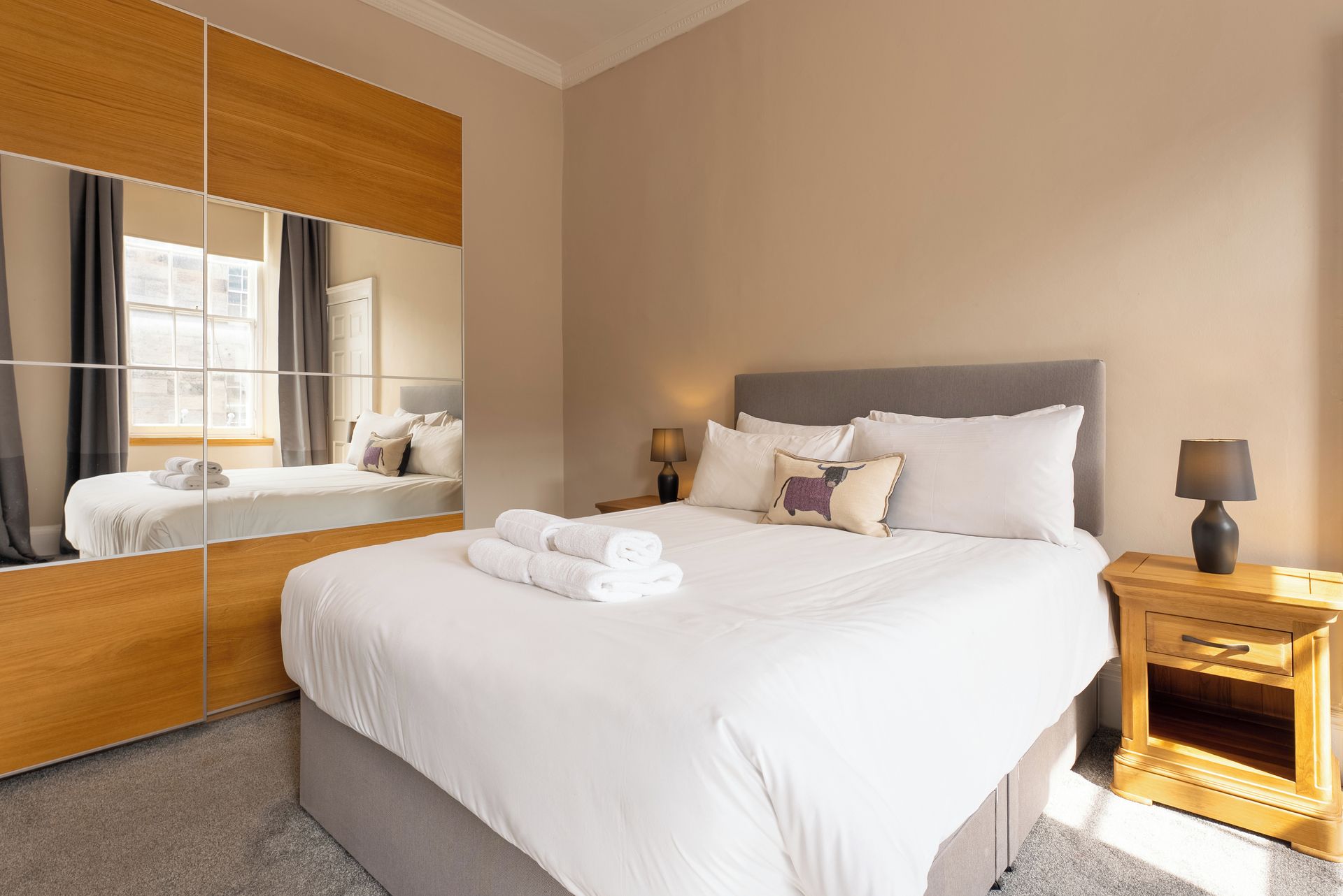 Habitación de hotel con una cama grande, una mesita de noche con lámpara, un espejo y vistas al exterior.