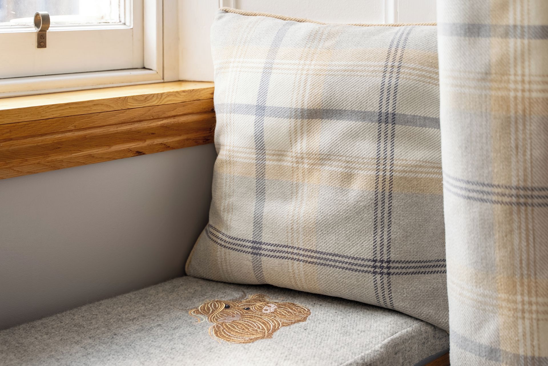 A corner of a room with a window, a wooden ledge, a cushion with a plaid pattern, and a cushion with a heart-shaped embroidery.
