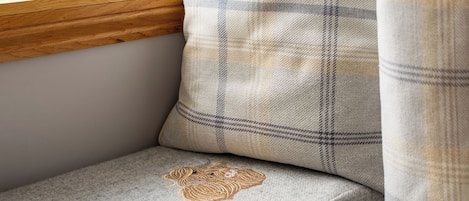 A corner of a room with a window, a wooden ledge, a cushion with a plaid pattern, and a cushion with a heart-shaped embroidery.