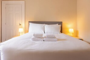 Ein ordentlich bezogenes Bett mit weißen Leinen und Handtüchern, zwei Nachttische mit Lampen und eine geschlossene Tür.