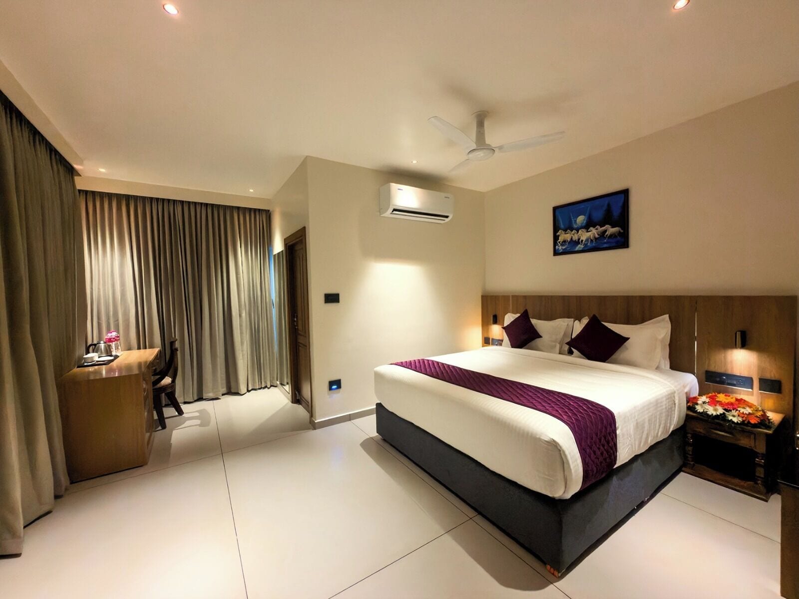 A hotel room with a large bed, a desk, a chair, a ceiling fan, and a painting on the wall.