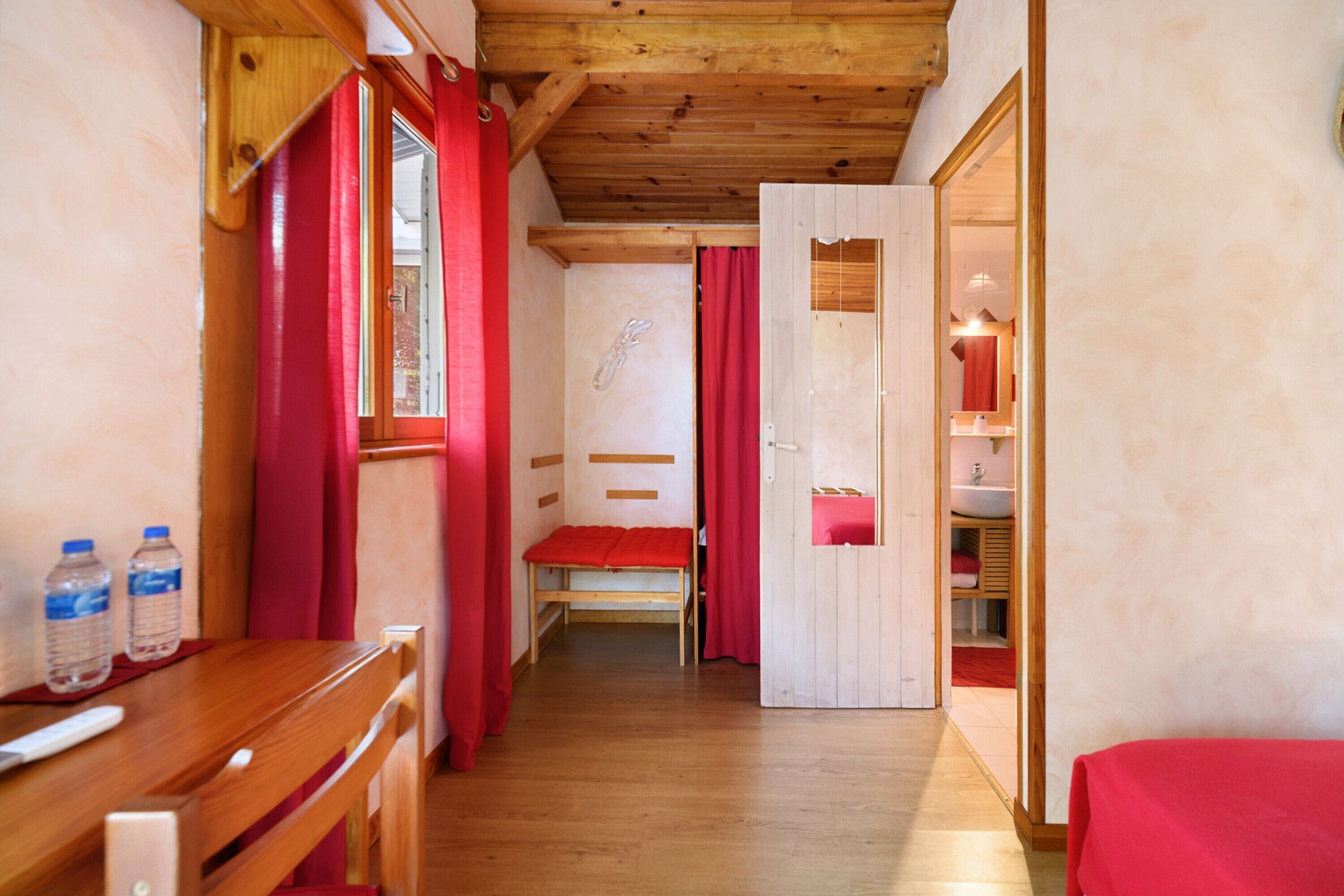 Un couloir étroit au plancher de bois, avec un banc, un lit recouvert d’une couverture rouge et une salle de bain visible à travers une porte ouverte.