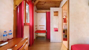 Un couloir étroit au plancher de bois, avec un banc, un lit recouvert d’une couverture rouge et une salle de bain visible à travers une porte ouverte.