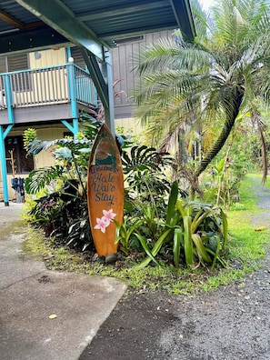 A wooden sign with 'Welcome to Wai'o Stay' written on it, placed in front of a building with a blue balcony and tropical plants.