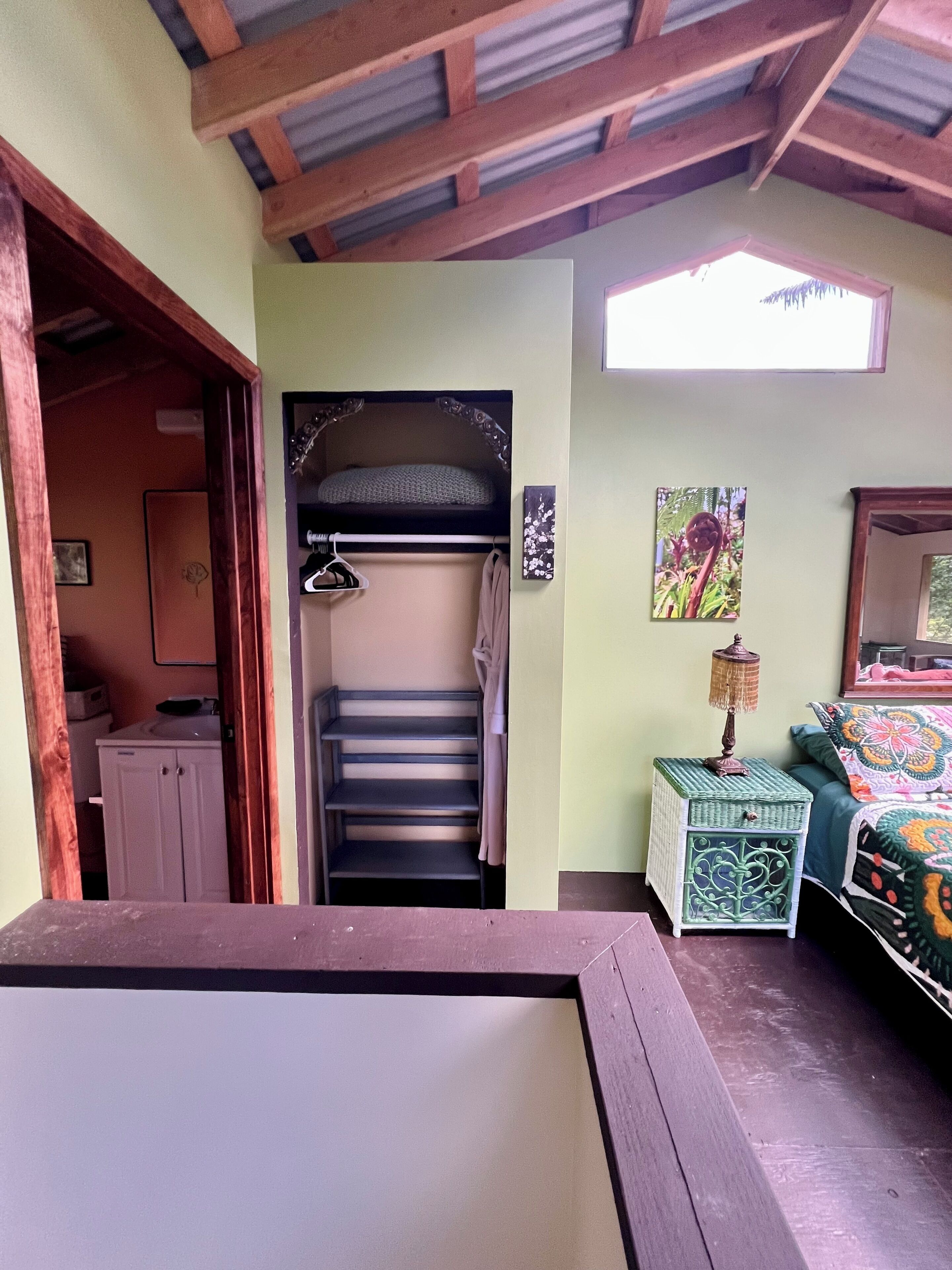 A bedroom with a bunk bed, a small table with a lamp, a bed with a colorful blanket, and a window with a view of greenery.